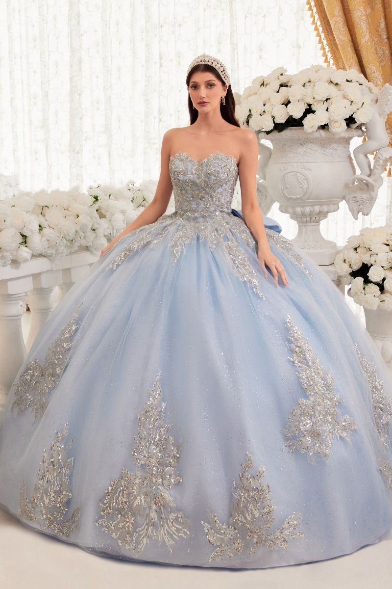 A woman stands indoors among large white floral arrangements, wearing the Ladivine Strapless Quinceanera Dress 15715—a light blue ball gown with silver embellishments.